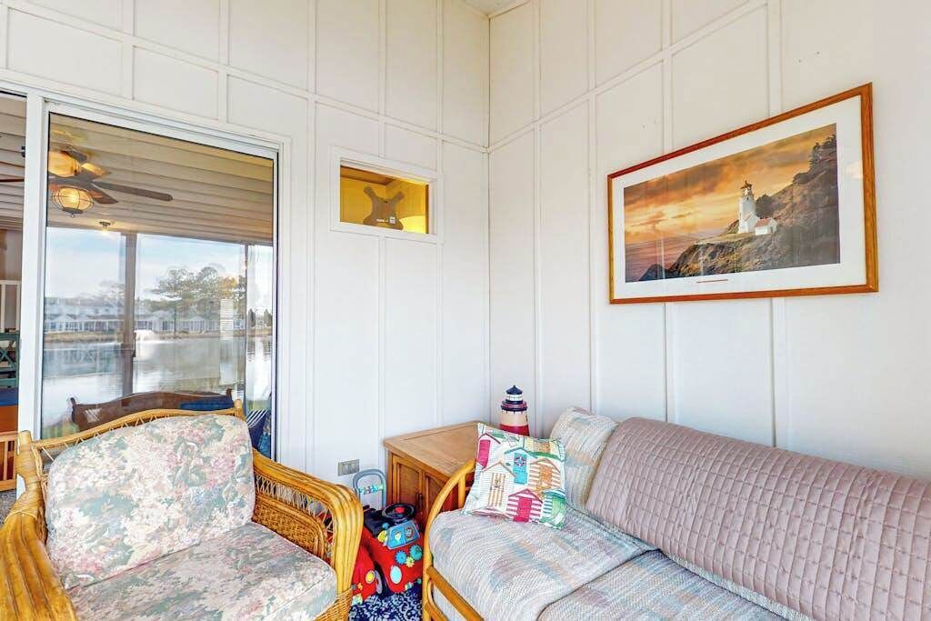 Et værelse med en fletstol, en polstret sofa, et lille bord med en lampe og et indrammet billede på væggen.