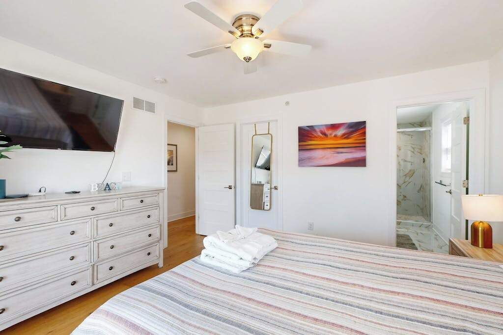 A bedroom with a bed, a dresser, a ceiling fan, a TV, a painting, and a mirror.