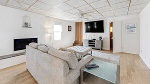 Ett vardagsrum med en soffa, ett soffbord i glas, en platt-TV, en öppen spis och en takfläkt.