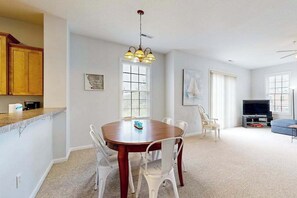 Ein Essbereich mit Holztisch und weißen Stühlen, eine Küche mit Holzschränken, einem Fernseher und einem gerahmten Bild an der Wand.