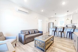 Una sala moderna con un sofá, una mesa de centro de vidrio y una zona de cocina con taburetes de bar.