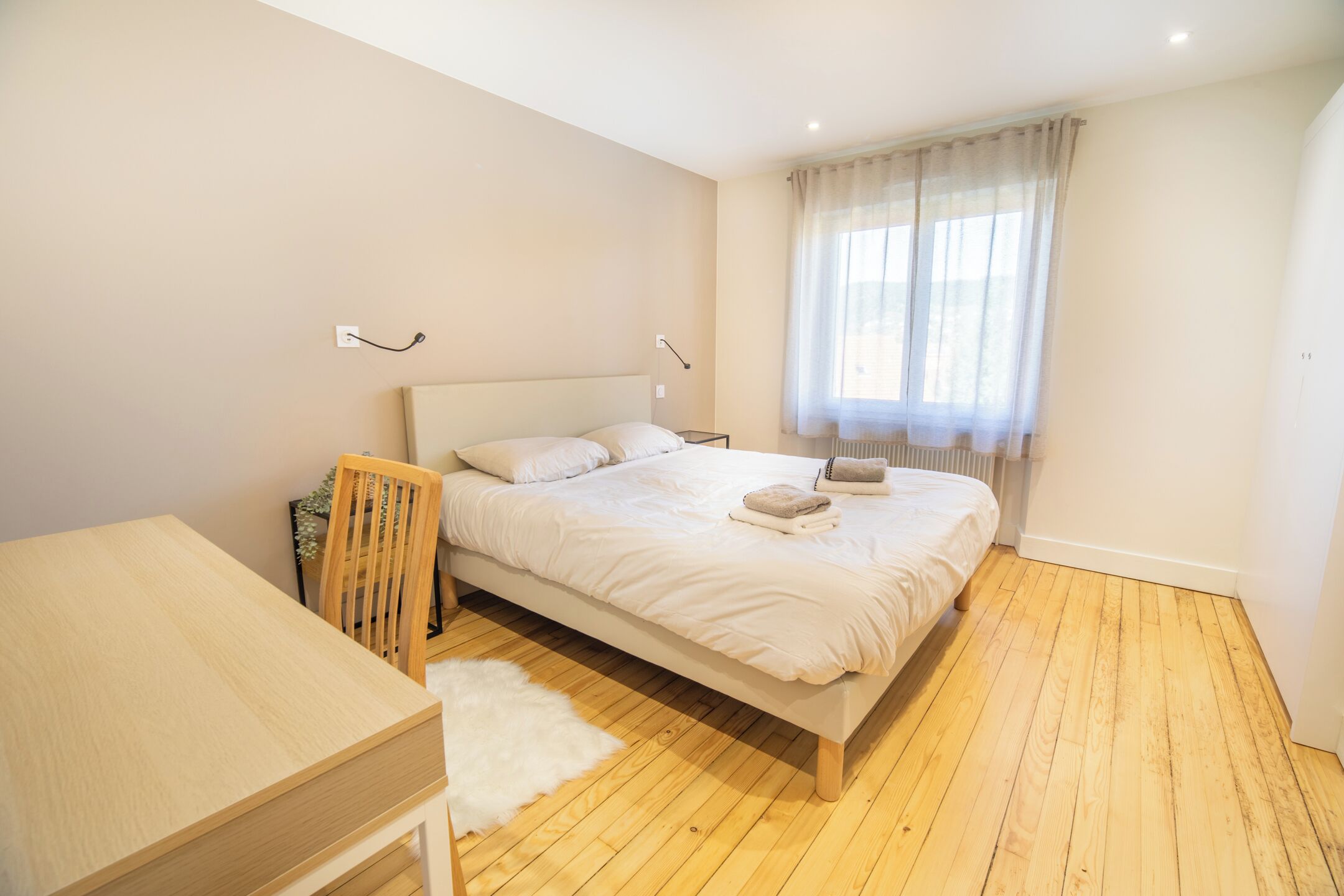 Une chambre à coucher avec un lit, un bureau en bois, une chaise, un tapis blanc et une fenêtre avec des rideaux.