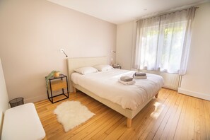 Une chambre à coucher avec un lit, une table de chevet, une petite étagère, un tapis blanc et une fenêtre avec des rideaux.