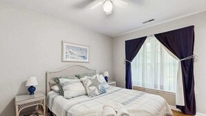 Een slaapkamer met een bed, een nachtkastje, een plafondventilator, een raam met gordijnen en een aan de muur bevestigd schilderij.