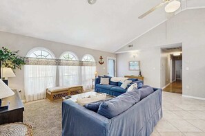 Un salón amplio con un sofá azul grande, una mesa de centro y una lámpara. Hay tres ventanas con cortinas, un ventilador de techo y una puerta que da a otra habitación.