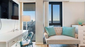Ein modernes Wohnzimmer mit Sofa, Wand-Fernseher, Schreibtisch mit Stuhl und großem Fenster.