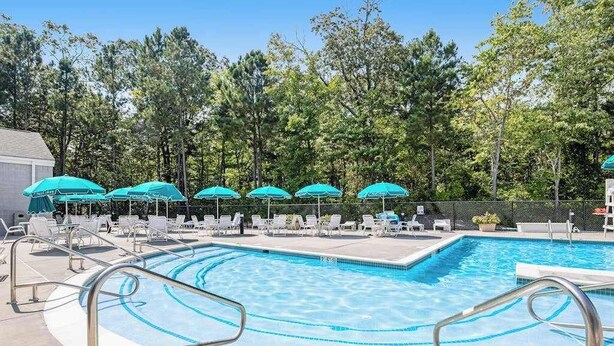 A swimming pool with lounge chairs and umbrellas, surrounded by trees. - Sea Colony Tennis condo with tennis court, pool, washer/dryer & covered porch (Bethany Beach)