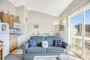 A compact living space with a kitchenette, a sofa with patterned cushions, a bed, and a large sliding glass door to a balcony.