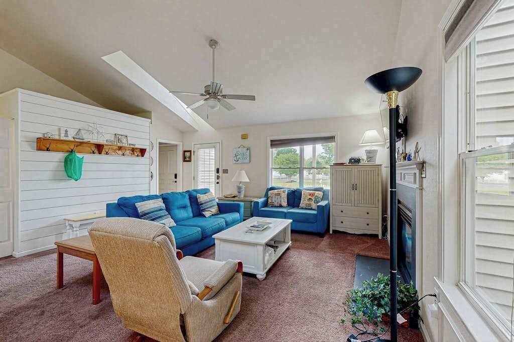 Una sala de estar con ventilador de techo, dos sofás azules, un sillón color beige, una mesa de centro blanca y una chimenea.