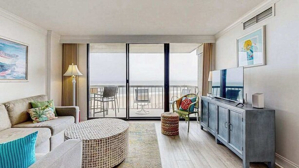 Habitació d'hotel moderna amb un balcó, un sofà, un otomà rodó, una cadira de vimetla, un armari i una televisió.