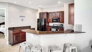 Una cocina moderna con armarios de madera oscura, un refrigerador negro, un microondas y una estufa. Hay dos taburetes blancos y un cartel en la pared.