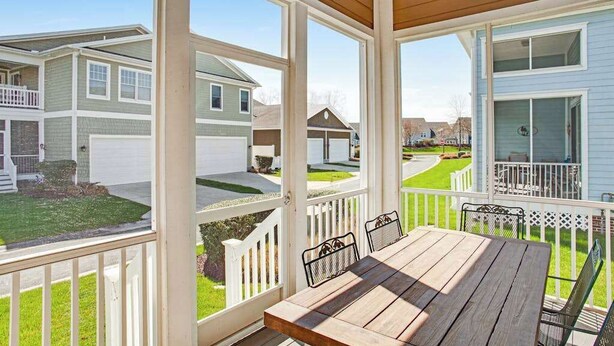 Un patio techado al aire libre con mesa y sillas de madera, rodeado de barandas blancas y vistas a casas vecinas.