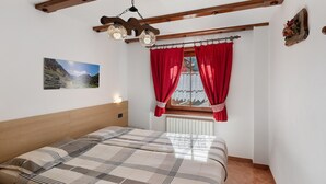 Ein Schlafzimmer mit einem Bett, einem Fenster mit roten Vorhängen, einer Holzbalkendecke und einer an der Wand befestigten Lampe.