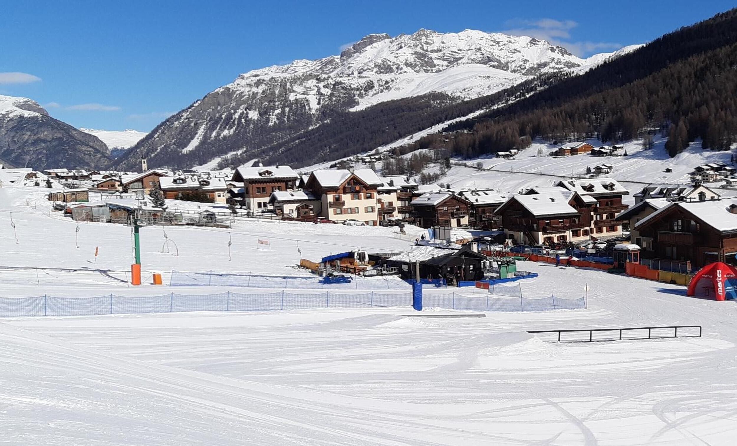 A snow-covered mountain village with chalet-style buildings, ski slopes, and a clear blue sky.