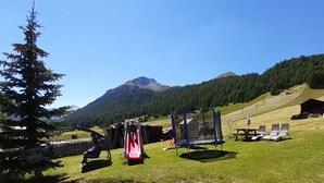 Ein Spielplatz mit Rutschen und Trampolin, umgeben von einer grünen Wiese mit einem Berg im Hintergrund.