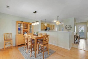 A dining area with a wooden table and chairs, a kitchen with wooden cabinets, pendant lighting, and a clock on the wall. - Sea Colony Condo with Fast WiFi & Gas Fireplace plus Shared Pools, Hot Tub, Gym (Bethany Beach)