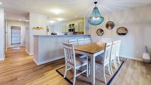 Un coin repas avec une table en bois et des chaises blanches, une cuisine avec des meubles de couleur claire et un luminaire au plafond.