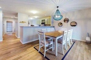 Ein Essbereich mit Holztisch und weißen Stühlen, eine Küche mit hellen Schränken und einer Deckenleuchte.