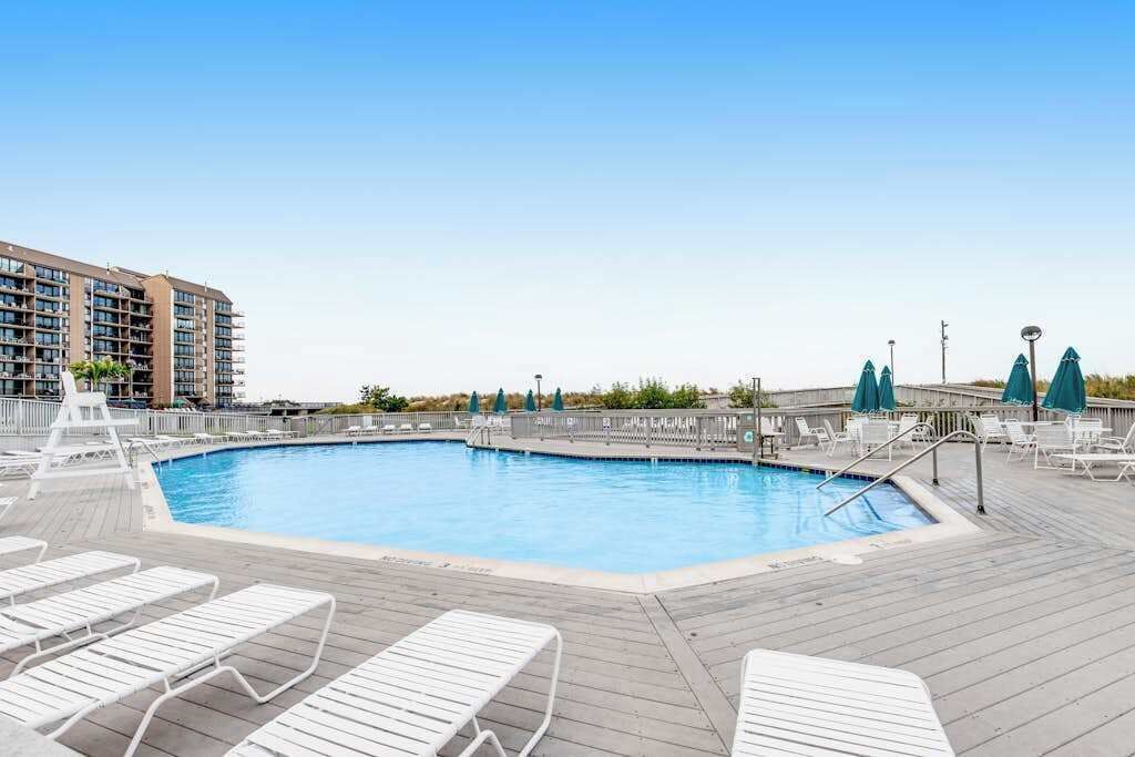 A swimming pool with lounge chairs, a wooden deck, and a hotel building in the background.