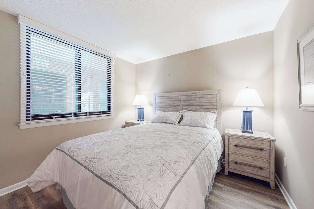 A bedroom with a bed, two bedside tables with lamps, a window with blinds, and a headboard.