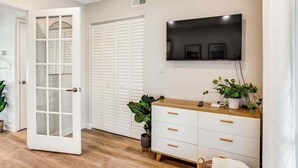 Une pièce avec une porte blanche, un placard blanc, une commode en bois, un téléviseur à écran plat et une plante en pot.