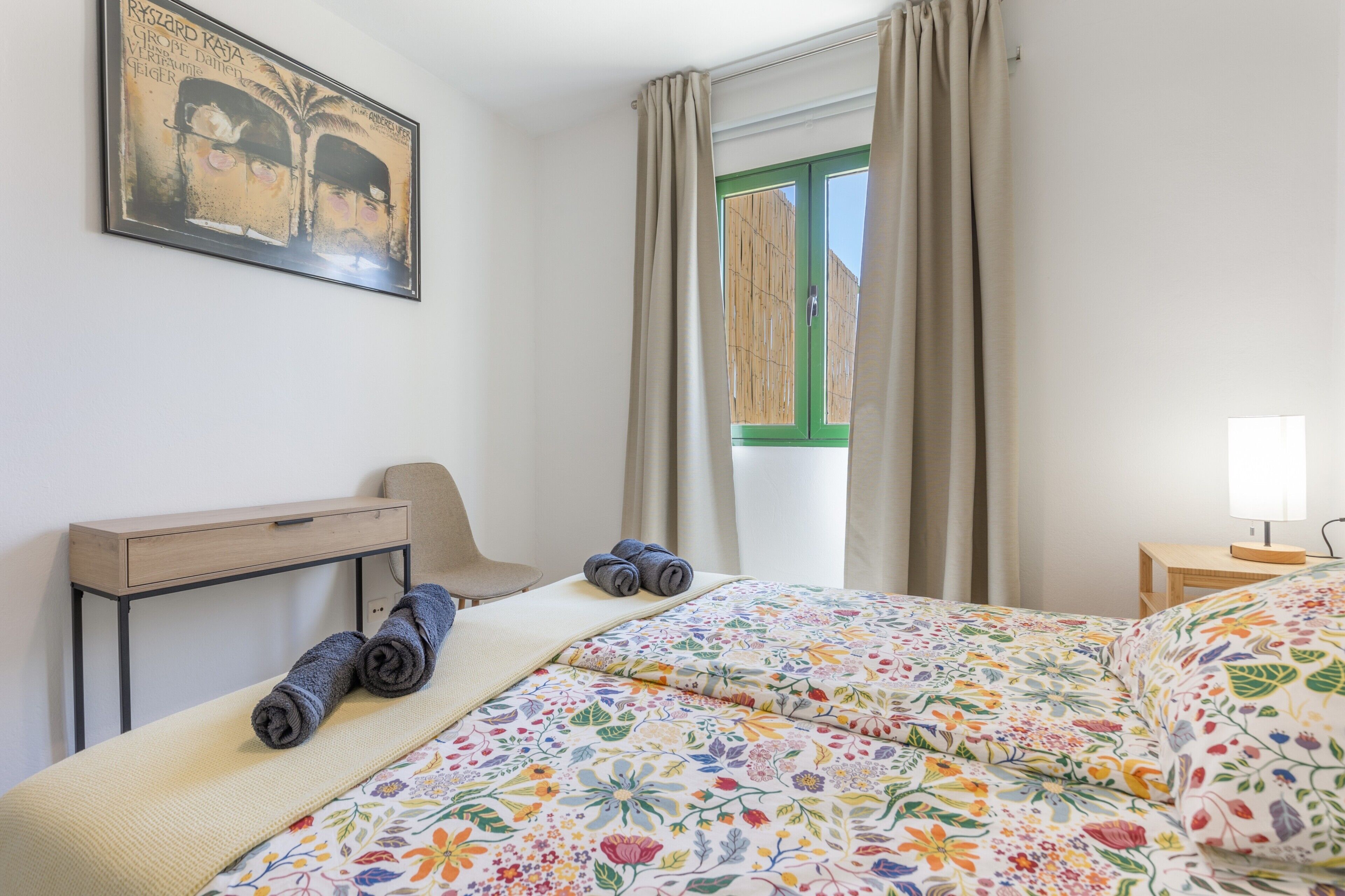 A bedroom with a bed, a desk, a chair, a window with curtains, and a painting on the wall.