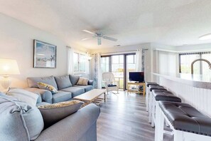 A modern living room with a sofa, coffee table, and a TV. There is a ceiling fan, a window with curtains, and a bench along the wall. - East of the Sun condo w/ central AC, W/D, & shared pool - under 1 mile to beach (Fenwick Island)