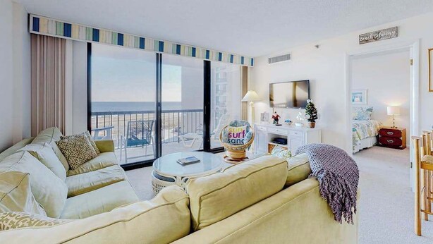 Quarto de hotel com sofá, mesa redonda, televisão, secretária pequena e vista para a praia.