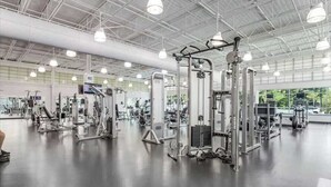 Uma academia ampla com diversos equipamentos de exercício, grandes janelas e pé direito alto.