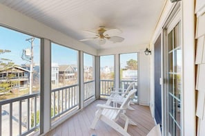 Eine überdachte Veranda mit weißen Stühlen, einem Deckenventilator und großen Fenstern mit Blick auf die Umgebung.