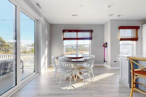 A modern kitchen with a round dining table, white chairs, and large windows offering a view of the outdoors. - Tidal Walk home with pond views, outdoor pool, decks, fast WiFi, & bikes (Ocean View)