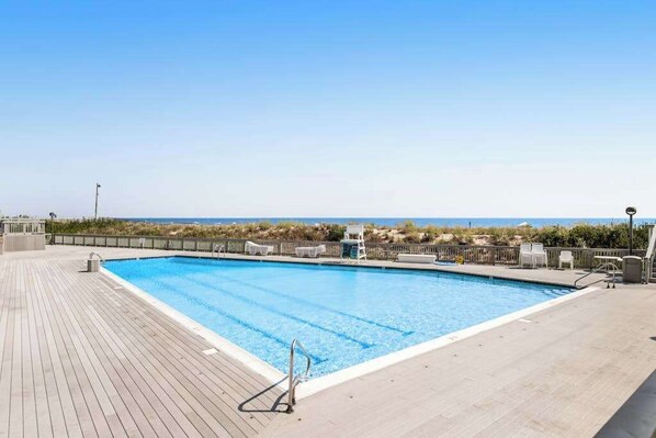Une piscine rectangulaire avec une terrasse en bois, entourée d’une clôture basse, et offrant une vue sur une plage avec des dunes.