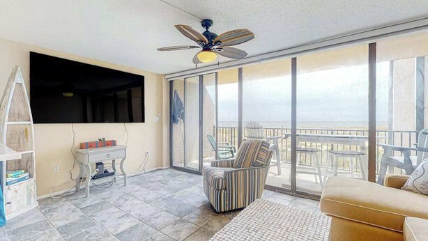 A spacious balcony with a view, a ceiling fan, a flat-screen TV, and a seating area with a sofa and chairs. - Sea Colony oceanfront condo with private balcony, shared pools, & gym (Bethany Beach)