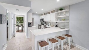 A modern kitchen with white cabinetry, a central island with stools, and a view to the outside. - Sea Colony condo with pools, hot tub, tennis, gym, & private deck (Bethany Beach)