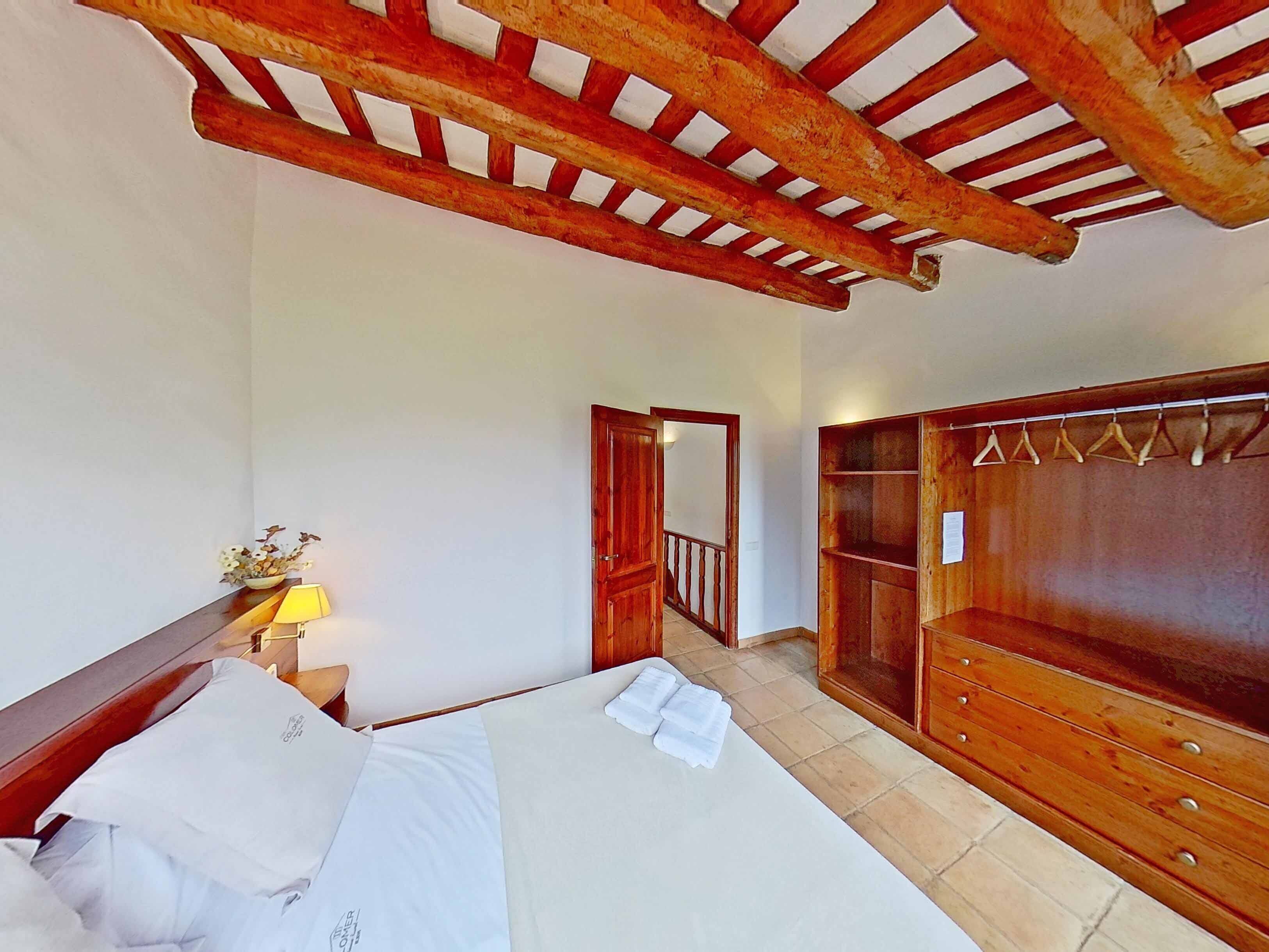 Een slaapkamer met een bed, nachtkastje, lamp, houten garderobe en een houten plafondconstructie.