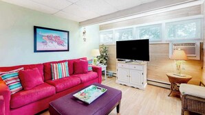 En stue med en rød sofa, et fladskærms-tv, et lille bord med en lampe og et vindue med rullegardiner.