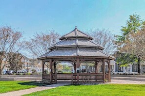 Ein Gazebo mit sechseckiger Dachform und offenen Seiten, umgeben von einer gepflasterten Fläche und Bäumen.
