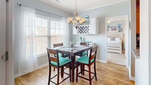 A dining area with a wooden table and chairs, a chandelier, and a view through a window with blinds. - Beautiful condo near ocean with pool, fast WiFi, central AC & washer/dryer (Lewes)