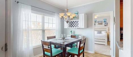 A dining area with a wooden table and chairs, a chandelier, and a view through a window with blinds.