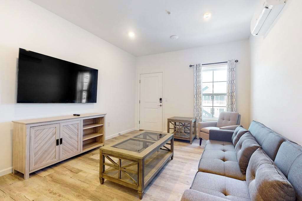 A living room with a flat-screen TV, wooden cabinet, glass coffee table, grey sofa, and a small side table.