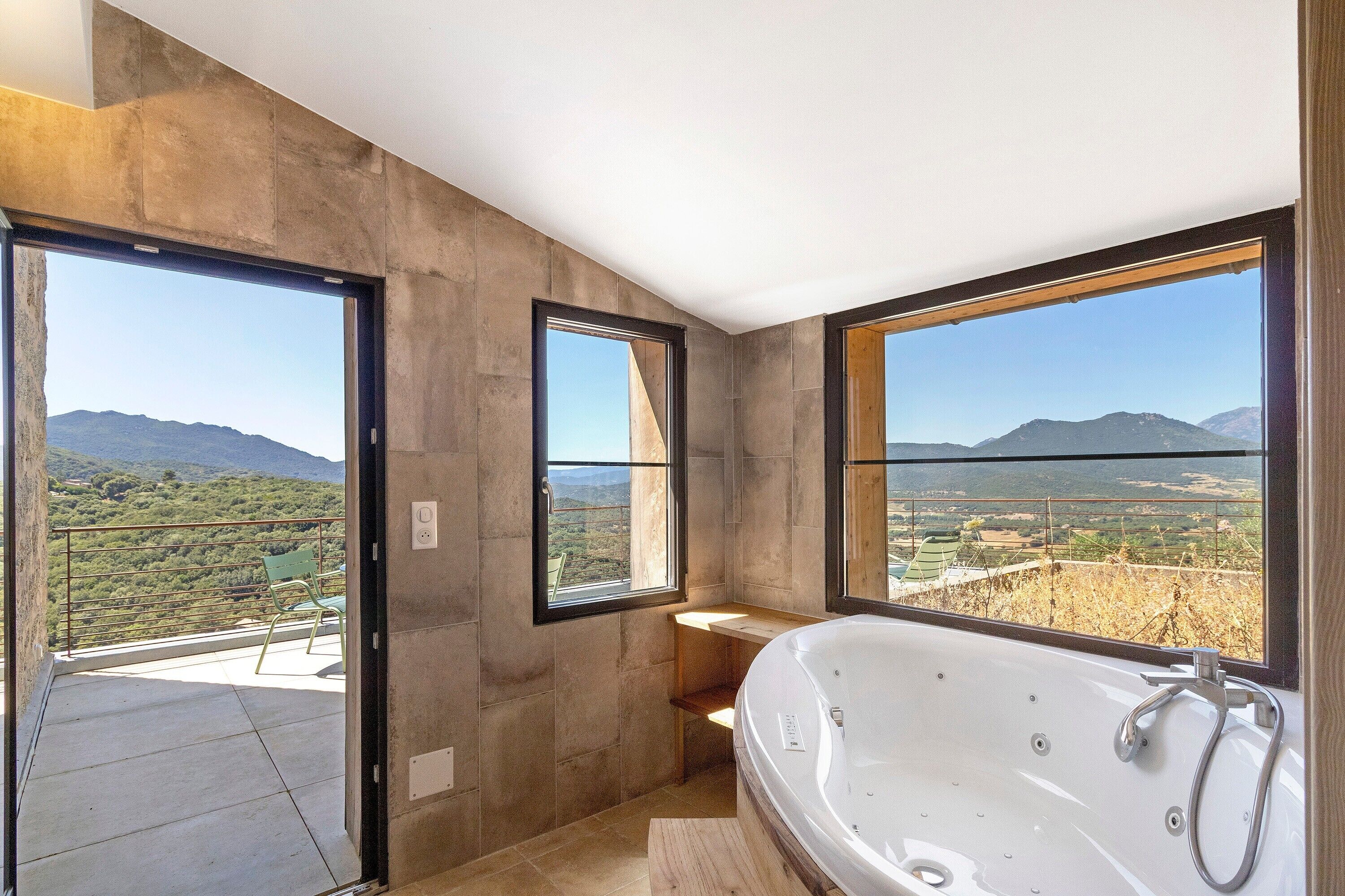 A modern bathroom with a large bathtub, two windows with mountain views, and a balcony with a chair.