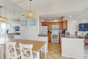A modern kitchen with white cabinets, a microwave, and a stove. There is a dining area with a wooden table and white chairs. - Sea Colony 7th floor condo with gym, shared sauna, and ocean views (Bethany Beach)