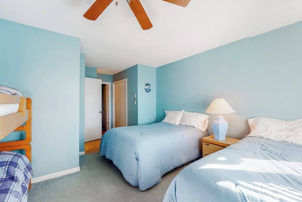 A bedroom with two beds, a ceiling fan, a nightstand with a lamp, and a door leading to another room.