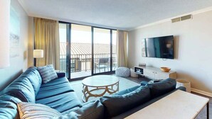 A modern living room with a sofa, a round coffee table, a flat-screen TV mounted on the wall, and a balcony with a view. - Sea Colony oceanfront 3rd floor condo w/ shared sauna, basketball court, balcony (Bethany Beach)