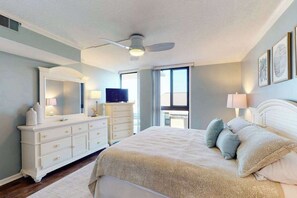 Ein Schlafzimmer mit Bett, Kommode, Schminktisch, Deckenventilator und großen Fenstern.