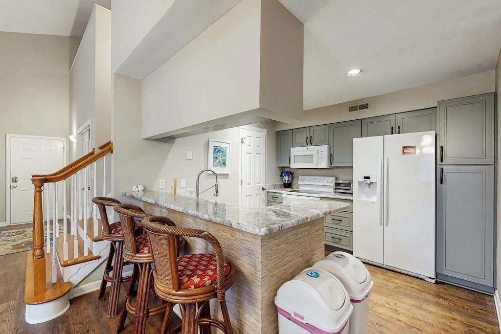 A kitchen with a marble island, wooden chairs, a microwave, a refrigerator, and a trash bin.