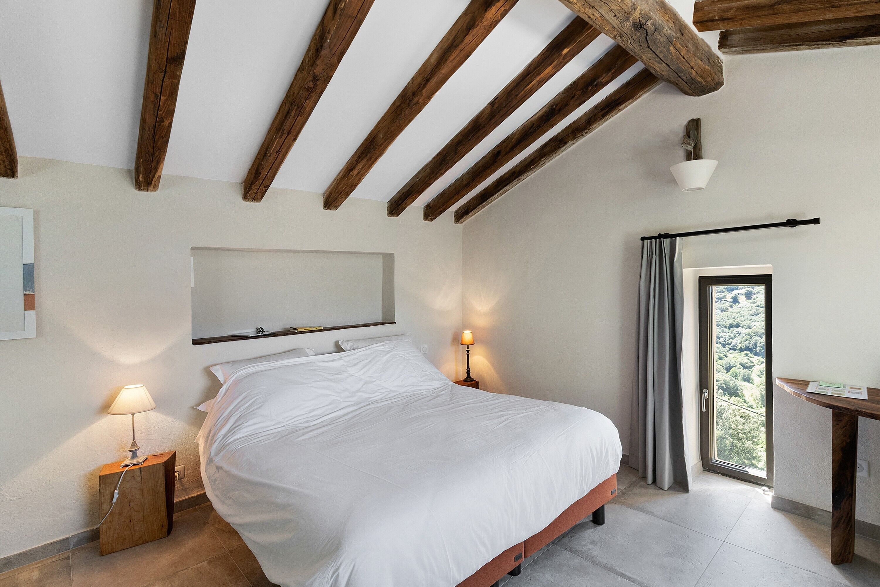 A bedroom with a bed, bedside lamps, a wooden ceiling, a door with a view, and a small table.