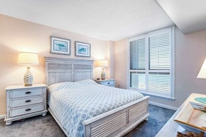 A bedroom with a bed, two bedside tables with lamps, a dresser, a window with blinds, and two framed pictures on the wall. - Sea Colony Oceanfront Condo w/Balcony, Free WiFi, Shared Pools, Hot Tub, Gym (Bethany Beach)