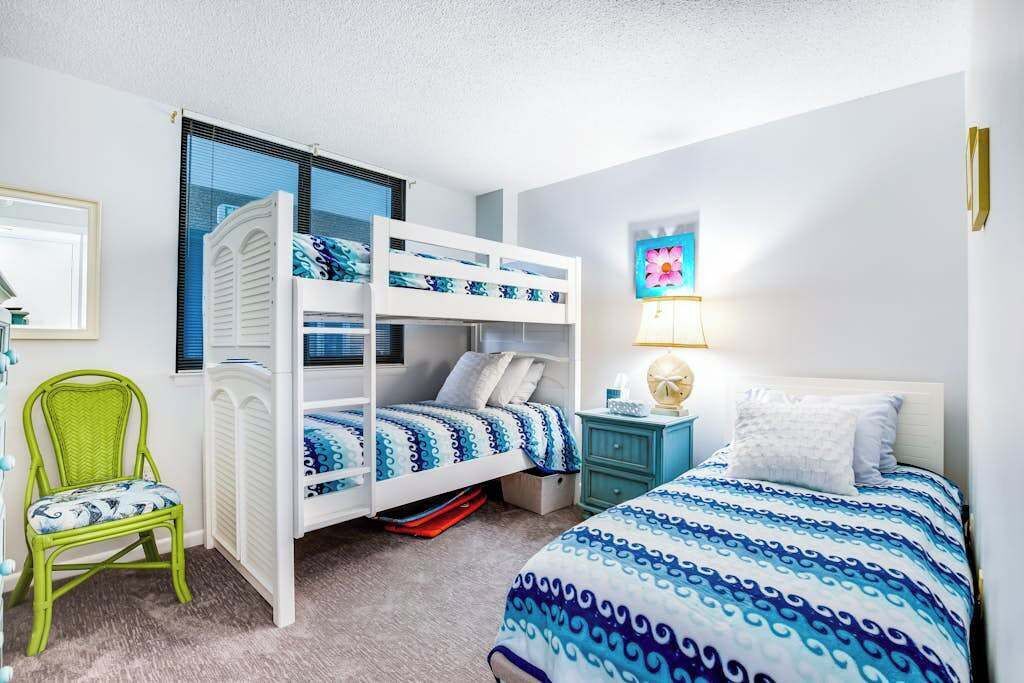 A room with bunk beds, a chair, a bedside table with a lamp, and a framed picture on the wall.
