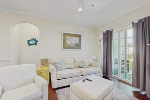 A living room with a sofa, armchair, and ottoman, a painting on the wall, and a large window with curtains.